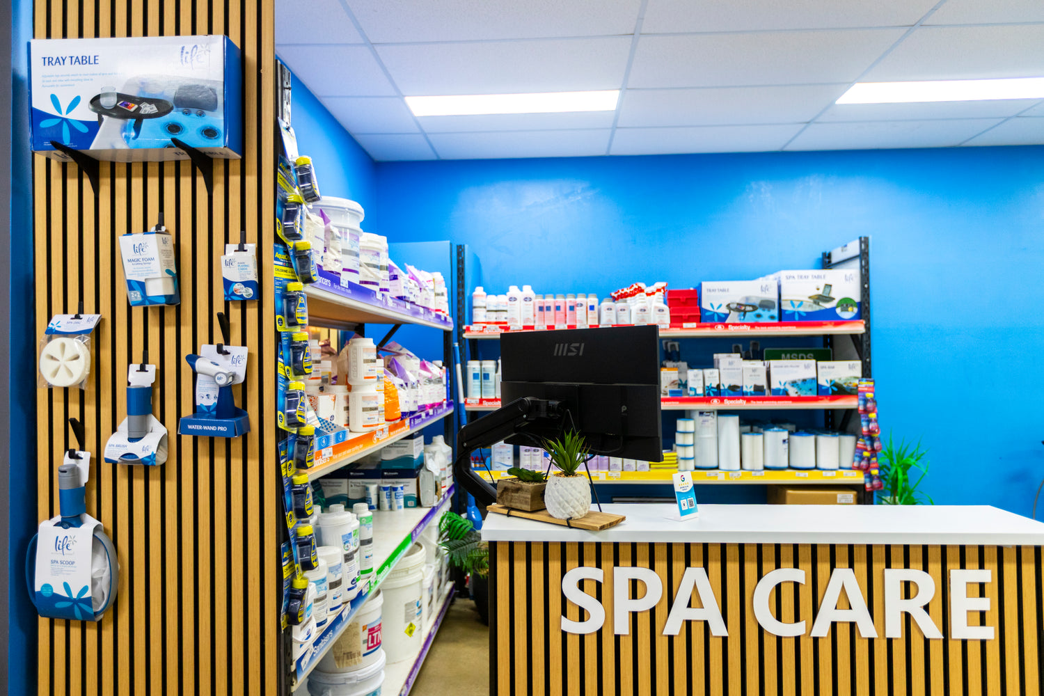A retail display area for spa care products, featuring a wooden slatted counter with 'SPA CARE' in white letters, shelves stocked with product boxes and bottles, and a bright blue wall in the background.